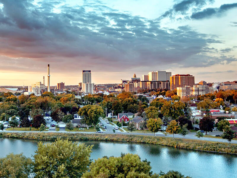 City skyline at sunset with river, trees, and buildings under a cloudy sky.