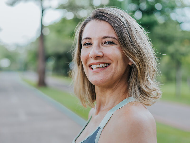 Smiling person outdoors, wearing a gray top with blurred greenery in the background.