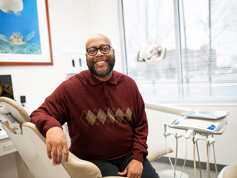 Person smiling while seated in a dental clinic, wearing a maroon sweater with argyle pattern.