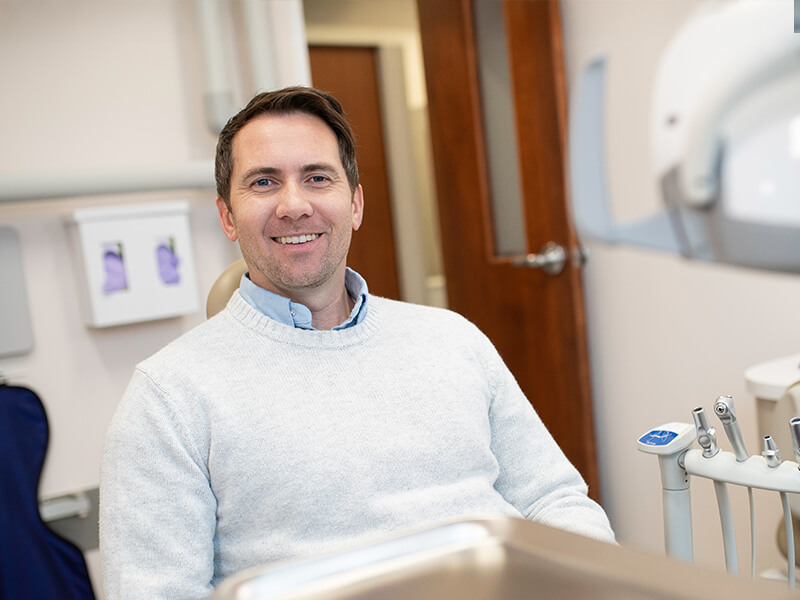 Smiling person in a dental office, wearing a light gray sweater, seated near dental equipment.