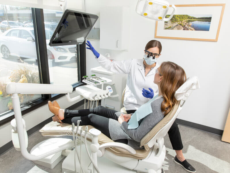Dentist in white coat and gloves discussing with patient in bright exam room.