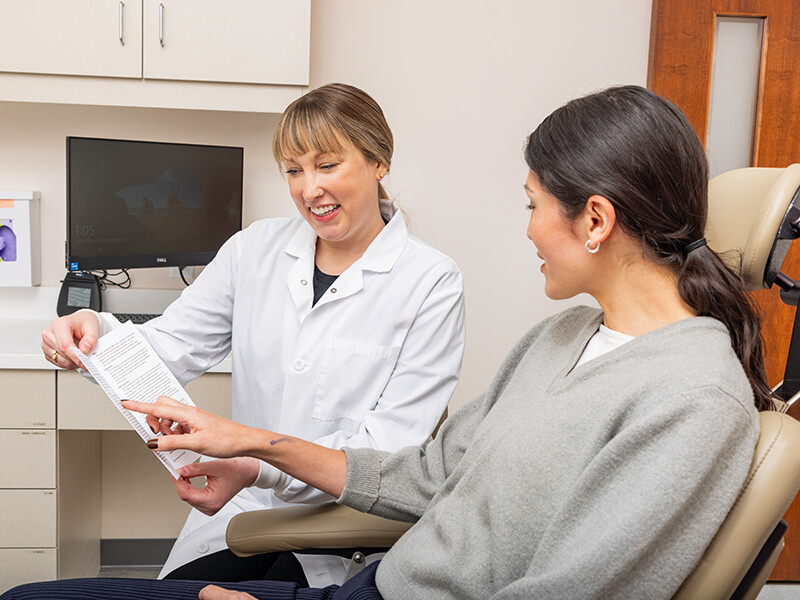 Dental professional in white coat showing paper to patient in grey sweater.