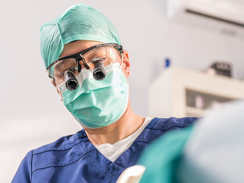 Dental professional in blue scrubs with magnifying loupes performs a procedure.