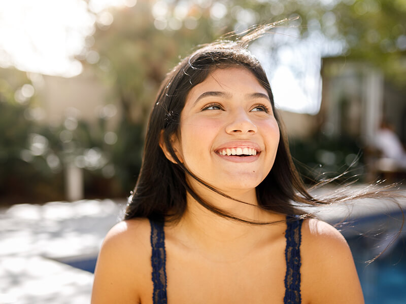 Person outdoors smiling, with long dark hair and a navy top, in a sunny setting.
