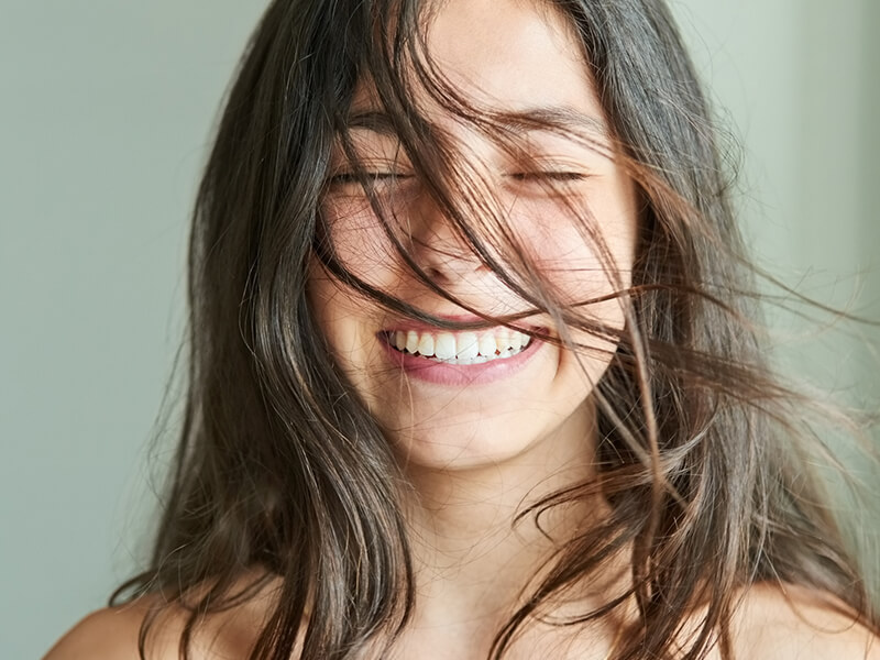 Person smiling with eyes closed, hair blowing across face, white teeth visible.