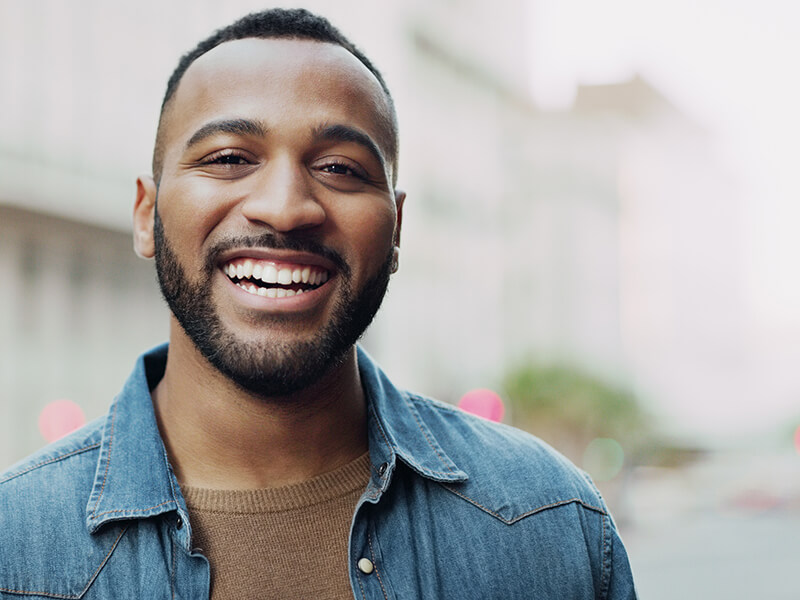 Smiling person with a beard, wearing a denim shirt and brown sweater outdoors.