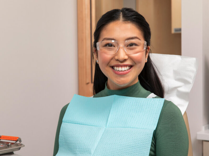 Person smiling in a dental chair, wearing protective glasses and a blue bib.